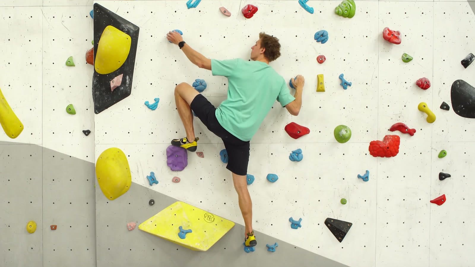 Alex Voit steigt an einer Boulderwand in der Halle auf einen hohen violetten Tritt, ein Rockover.